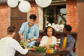 Multi-ethnic group of friends sitting at wooden table while enjoying dinner outdoors at Summer party, copy space