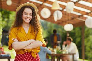 Colorful waist up portrait of smiling young woman  while posing outdoors in Summer with friends and family enjoying dinner at terrace in background, copy space