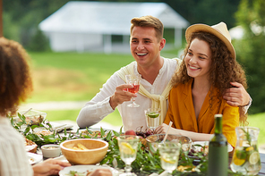 Portrait of young couple embracing while sitting table holding drinks and enjoying dinner with friends outdoors at Summer party, copy space