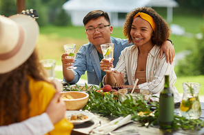 Portrait of mixed-race young couple embracing while sitting table holding drinks and enjoying dinner with friends outdoors at Summer party, copy space