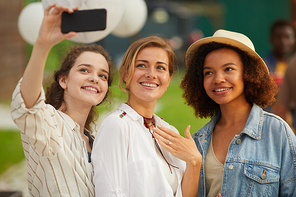 Waist up portrait of three beautiful young women taking selfie photo via smartphone during outdoor party in Summer