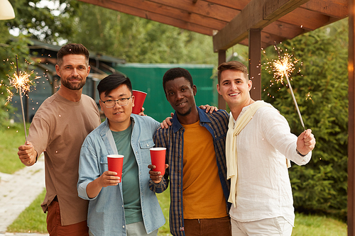 Multi-ethnic group of young men holding sparklers and smiling at camera while enjoying Summer party at outdoor terrace, copy space