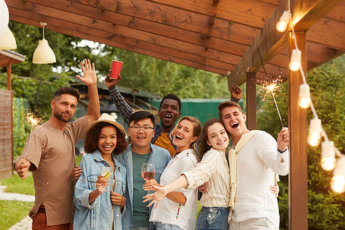 Multi-ethnic group of carefree young people holding sparklers and smiling at camera while enjoying Summer party at outdoor terrace, copy space