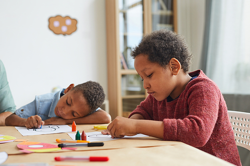 Side view portrait of African-American boy drawing pictures with crayons while enjoying art class in preschool, copy space