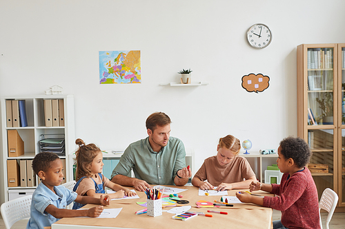 High angle view at male teacher working with multi-ethnic group of children drawing pictures during art class in school or development center, copy space