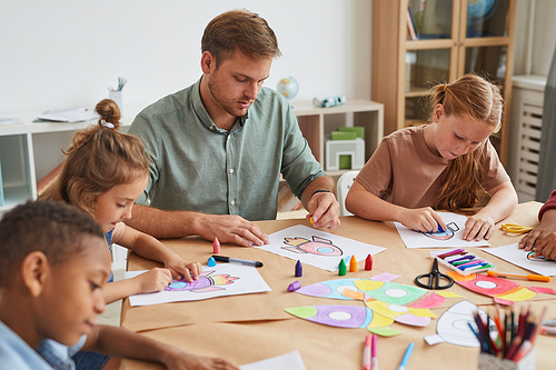 Portrait of male teacher working with multi-ethnic group of children drawing pictures during art class in school or development center, copy space