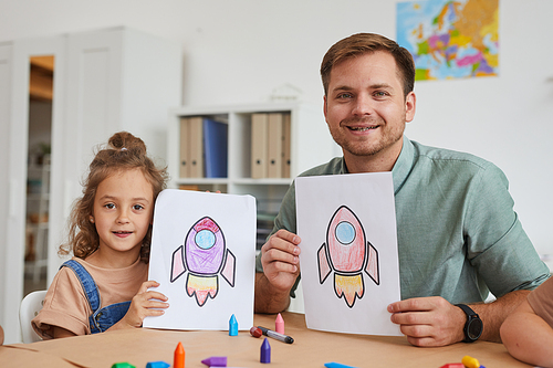 Portrait of smiling young man and little girl showing pictures of space rockets while enjoying art class in preschool together, copy space