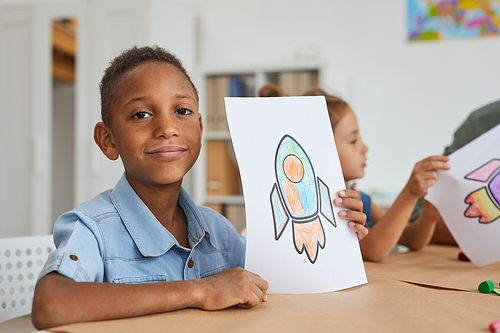 Portrait of cute African-American boy showing pictures of space rocket and smiling at camera while enjoying art class in pre school or children development center, copy space