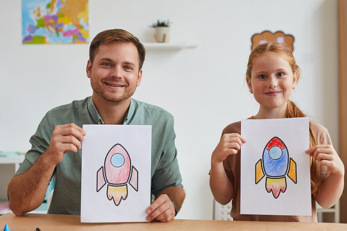 Portrait of smiling father and daughter showing pictures of space rockets while enjoying art class in school or development center together, copy space