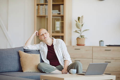 Portrait of bald adult woman looking away pensively while sitting on couch in modern home interior, alopecia and cancer awareness, copy space