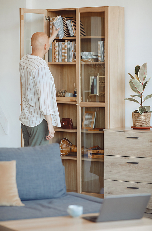 Vertical back view at bald person putting book on shelf on wooden bookcase in modern home interior, alopecia and cancer awareness, copy space