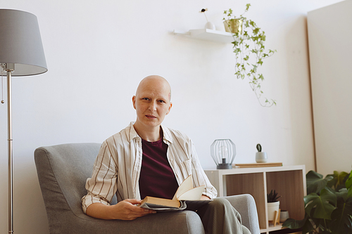 Portrait of bald mature woman looking at camera while reading book sitting in comfortable armchair at home, alopecia and cancer awareness, copy space