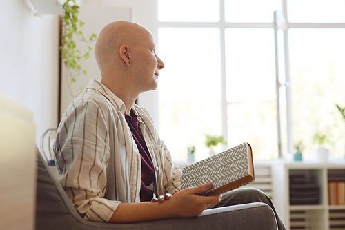 Minimal side view portrait of bald young woman holding book and looking away pensively while sitting in armchair at home, copy space