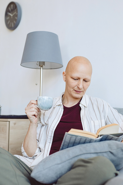 Vertical portrait of bald mature woman reading book sitting in comfortable armchair at home and enjoying cup of coffee, alopecia and cancer awareness, copy space
