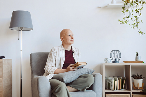 Minimal portrait of bald adult woman holding book and looking away pensively while sitting in comfy armchair at home, alopecia and cancer awareness, copy space