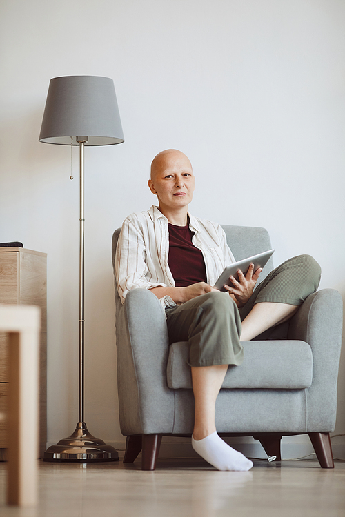Minimal full length portrait of bald adult woman holding book and looking at camera pensively while sitting in comfy armchair at home, alopecia and cancer awareness