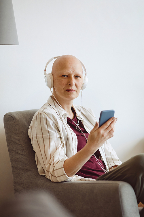 Minimal portrait of bald adult woman looking at camera while listening to music via smartphone at home, alopecia and cancer awareness, copy space