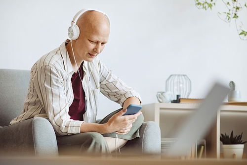 Minimal side view portrait of bald adult woman wearing headphones while listening to music via smartphone in cozy home interior, alopecia and cancer awareness, copy space
