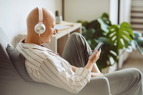Minimal back view portrait of bald adult woman wearing headphones while listening to music via smartphone in modern home interior, alopecia and cancer awareness, copy space