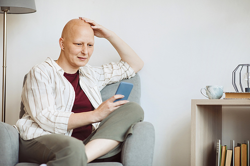Minimal portrait of bald adult woman using smartphone and smiling while sitting in cozy armchair at home, alopecia and cancer awareness