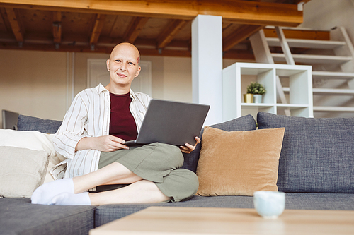 Full length portrait of bald adult woman working from home and looking at camera while sitting on comfy couch in modern home interior, alopecia and cancer awareness, copy space