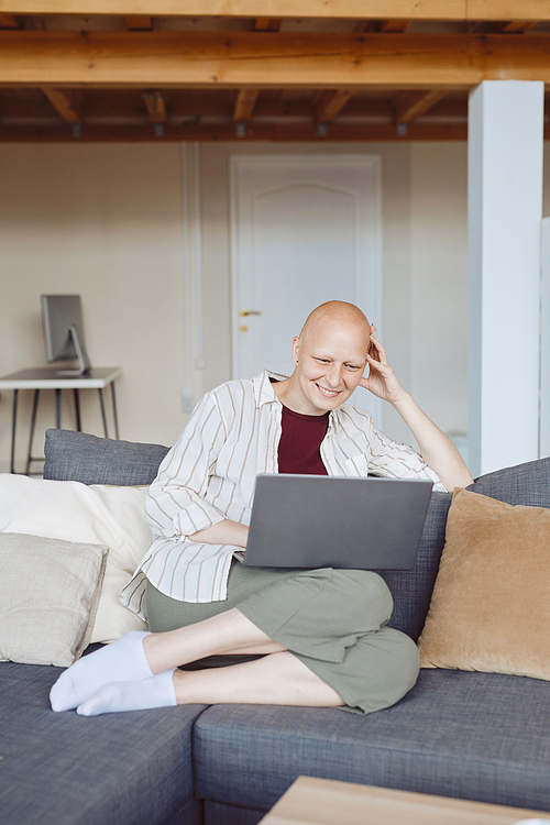 Vertical full length portrait of bald adult woman smiling happily looking at laptop screen while sitting on comfy couch at home, alopecia and cancer awareness, copy space