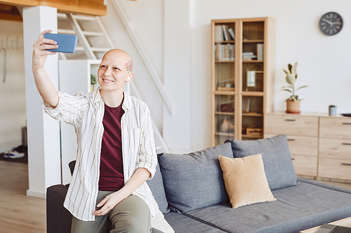 Minimal portrait of bald adult woman with head tattoo taking selfie and smiling happily while standing in modern home interior, alopecia and cancer awareness, copy space