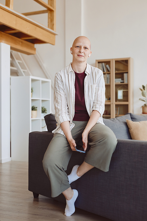 Vertical full length portrait of bald adult woman looking at camera while relaxing casually at home in modern interior, alopecia and cancer awareness, copy space