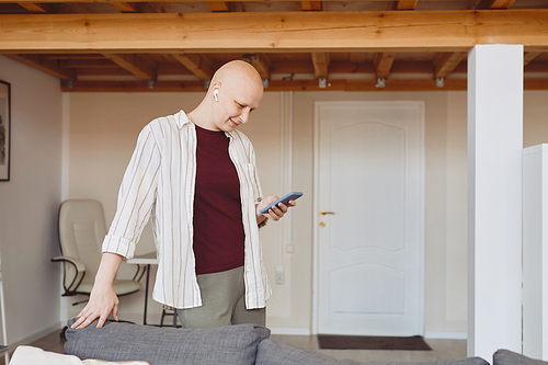 Side view portrait of smiling bald woman using smartphone with wireless earphones while enjoying music at home in modern interior, alopecia and cancer awareness, copy space