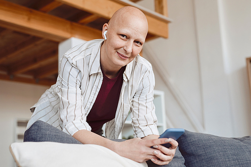 Portrait of smiling adult woman looking at camera and holding smartphone while leaning on couch at home in modern interior, alopecia and cancer awareness, copy space