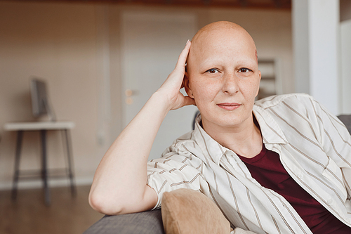 Minimal portrait of bald adult woman looking at camera while sitting on couch in warm-toned home interior, alopecia and cancer awareness, copy space