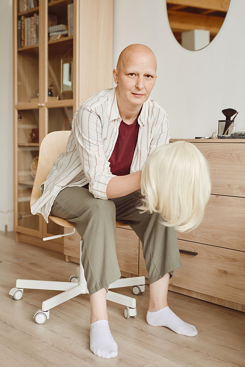 Vertical full length portrait of bald woman holding wig while sitting by mirror and looking at camera in minimal home interior, alopecia and cancer awareness