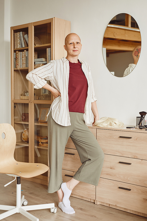 Vertical full length portrait of confident bald woman standing by mirror and looking at camera while posing in minimal home interior, alopecia and cancer awareness