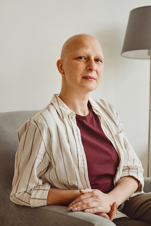 Vertical warm-toned portrait of confident bald woman looking at camera while sitting on couch in minimal home interior, alopecia and cancer awareness