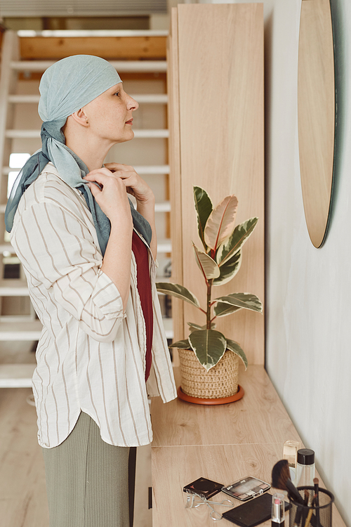 Vertical side view portrait of modern bald woman putting on head scarf while looking in mirror standing in home interior, alopecia and cancer awareness