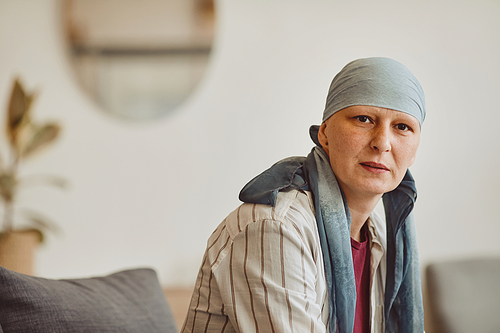 Warm-toned portrait of bald adult woman wearing headscarf looking at camera while posing in minimal home interior, alopecia and cancer awareness, copy space