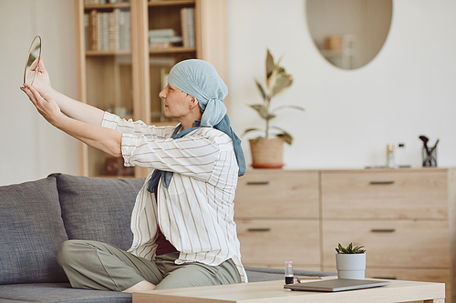 Warm-toned portrait of bald woman wearing headscarf looking in mirror while sitting on couch in cozy home interior, copy space
