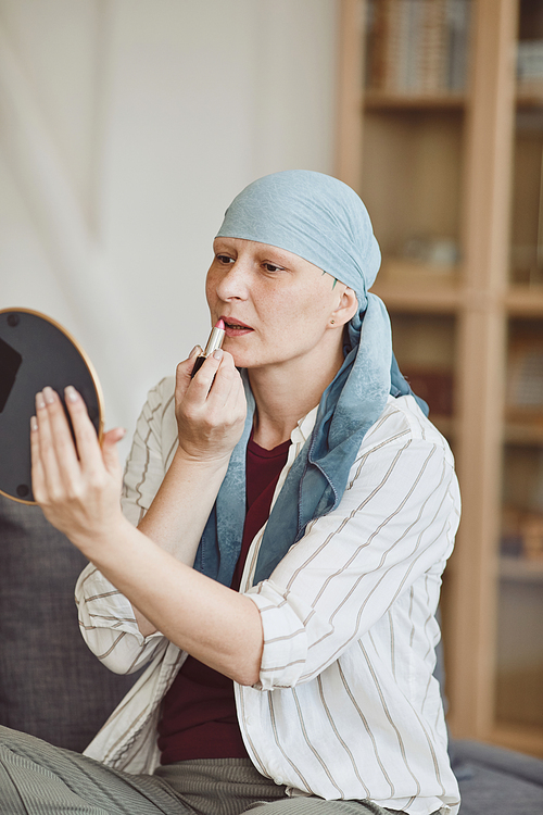 Vertical warm-toned portrait of confident bald woman putting on makeup and lipstick while looking in mirror at home, embracing beauty, alopecia and cancer awareness
