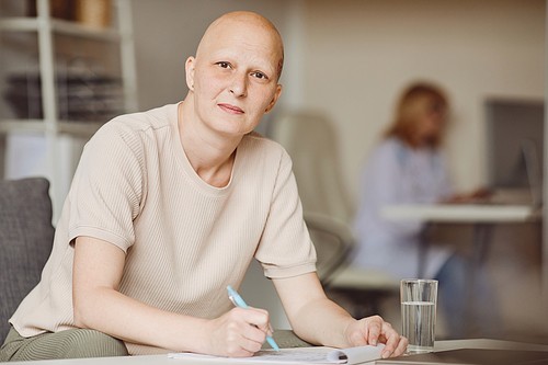 Warm toned portrait of adult bald woman looking at camera while filling in papers at doctors office during alopecia and cancer recovery, copy space