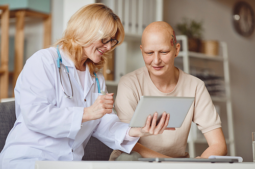 Warm-toned portrait of bald woman looking at digital tablet during consultation with female doctor on alopecia and cancer recovery
