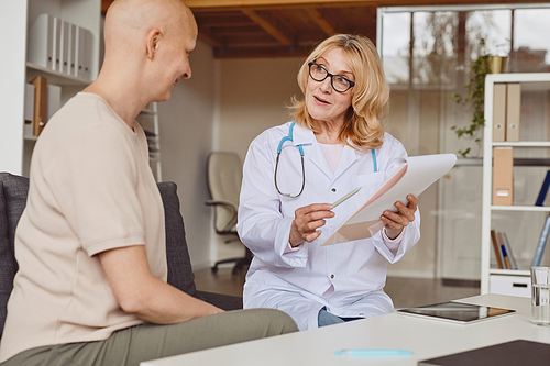 Warm-toned portrait of cheerful female doctor holding clipboard and talking to bald patient during consultation on alopecia and cancer recovery, copy space