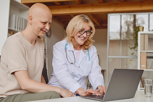 Warm-toned portrait of cheerful female doctor using laptop and talking to bald patient during consultation on alopecia and cancer recovery, copy space