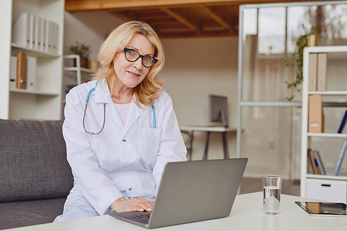 Warm-toned portrait of cheerful female doctor using laptop and smiling at camera while working in modern clinic, copy space