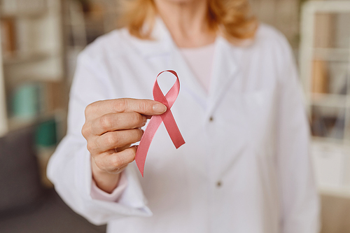 Warm-toned close up of unrecognizable female doctor holding pink ribbon as symbol of breast cancer awareness, copy space