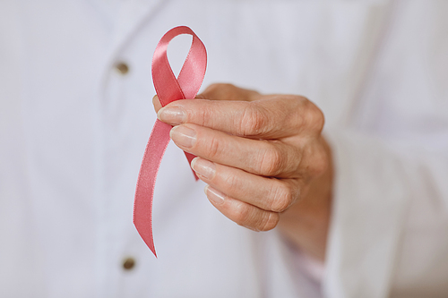Extreme close up of unrecognizable female doctor holding pink ribbon against white lab coat as symbol of breast cancer awareness, copy space