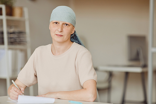 Warm-toned waist up portrait of mature bald woman wearing headscarf looking at camera while filling papers in doctors office, alopecia and cancer awareness, copy space