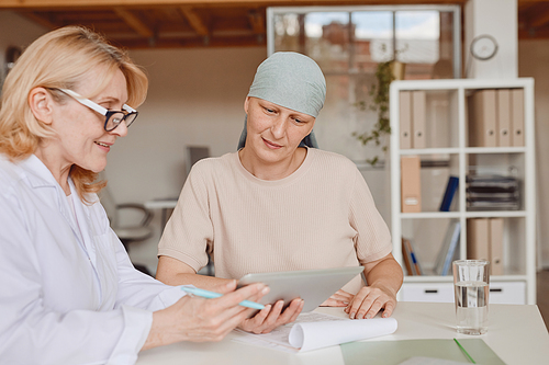 Warm-toned portrait of mature bald woman listening to female doctor showing info at digital tablet during consultation on alopecia and cancer recovery, copy space