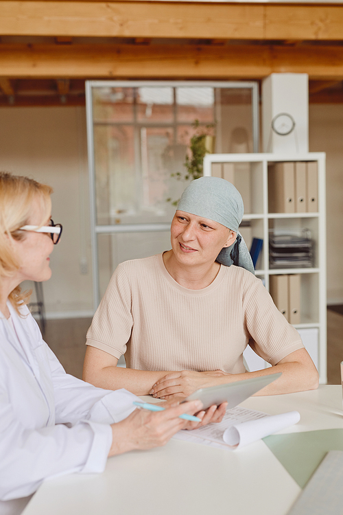 Vertical warm-toned portrait of smiling bald woman listening to female doctor showing info at digital tablet during consultation on alopecia and cancer recovery