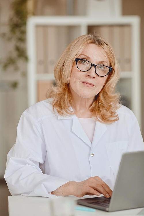 Vertical warm-toned portrait of mature female doctor looking at camera while working at desk in modern clinic office