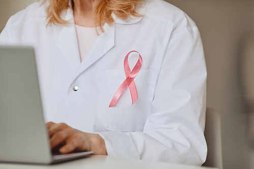 Close up background of pink ribbon pinned on white lab coat of female doctor using laptop, breast cancer awareness symbol, copy space